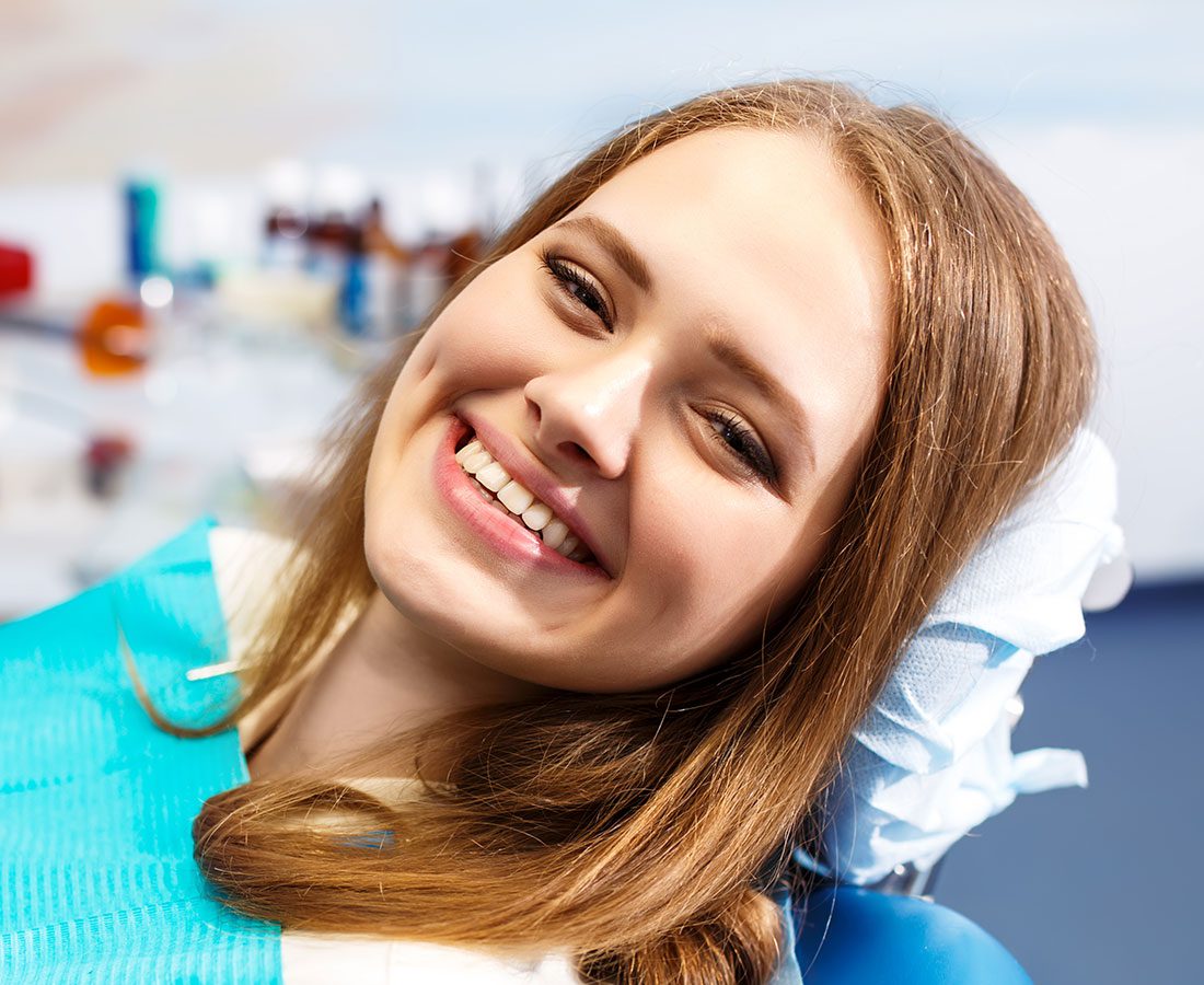 A young woman with long brown hair smiles while reclining in a dentist’s chair, wearing a blue dental bib. The background shows blurred dental equipment.