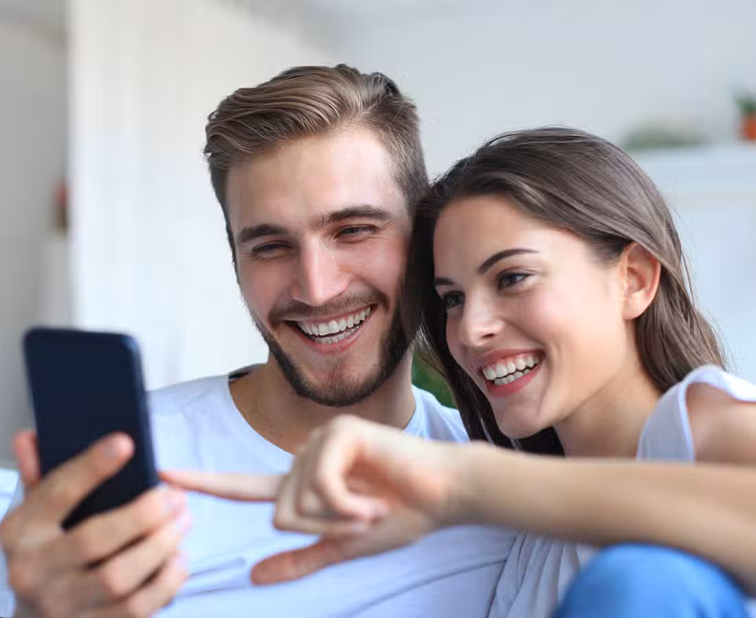A smiling man and woman sit close together, looking at a smartphone. The woman points at the screen, and both appear happy and engaged. They are indoors, wearing casual white shirts.